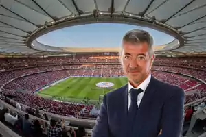 Un hombre de traje posando frente a un estadio de fútbol lleno de espectadores.