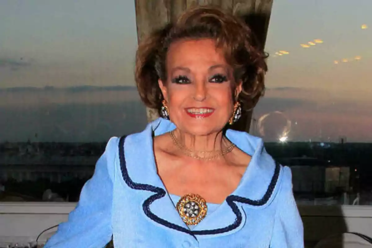Mujer mayor sonriendo con un traje azul claro y joyas elegantes, con un fondo de cielo al atardecer.