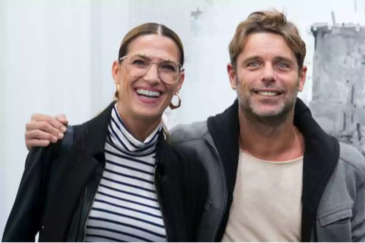Dos personas sonriendo y posando para la cámara, una mujer con gafas y una camiseta de rayas y un hombre con una camiseta clara y una chaqueta gris.