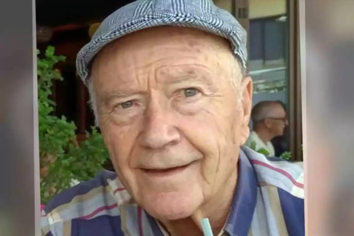 Un hombre mayor con una gorra a cuadros y una camisa de rayas sonríe mientras sostiene una pajilla en la boca, con un fondo de plantas y una ventana.