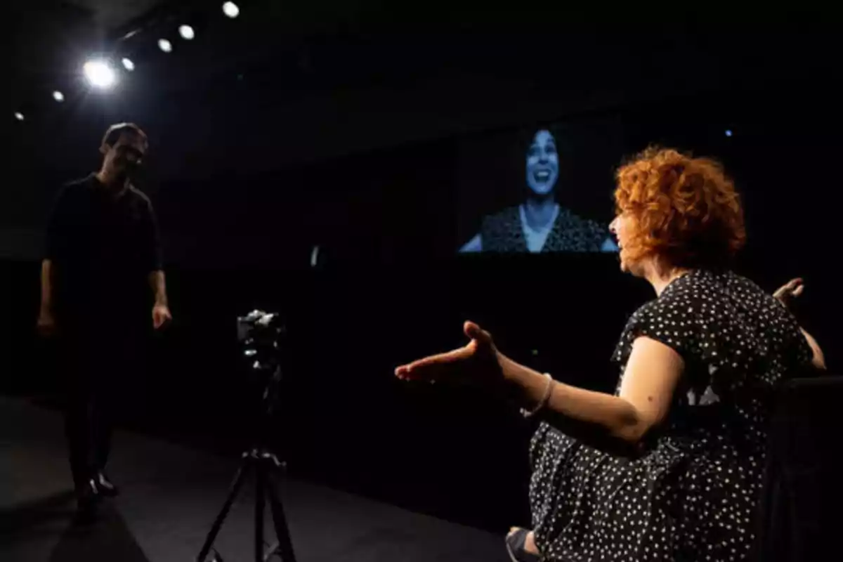 Una mujer pelirroja con un vestido de lunares está sentada y gesticulando mientras un hombre se acerca a ella en un escenario oscuro iluminado por focos, con una cámara en un trípode y una pantalla en el fondo mostrando la imagen de la mujer.