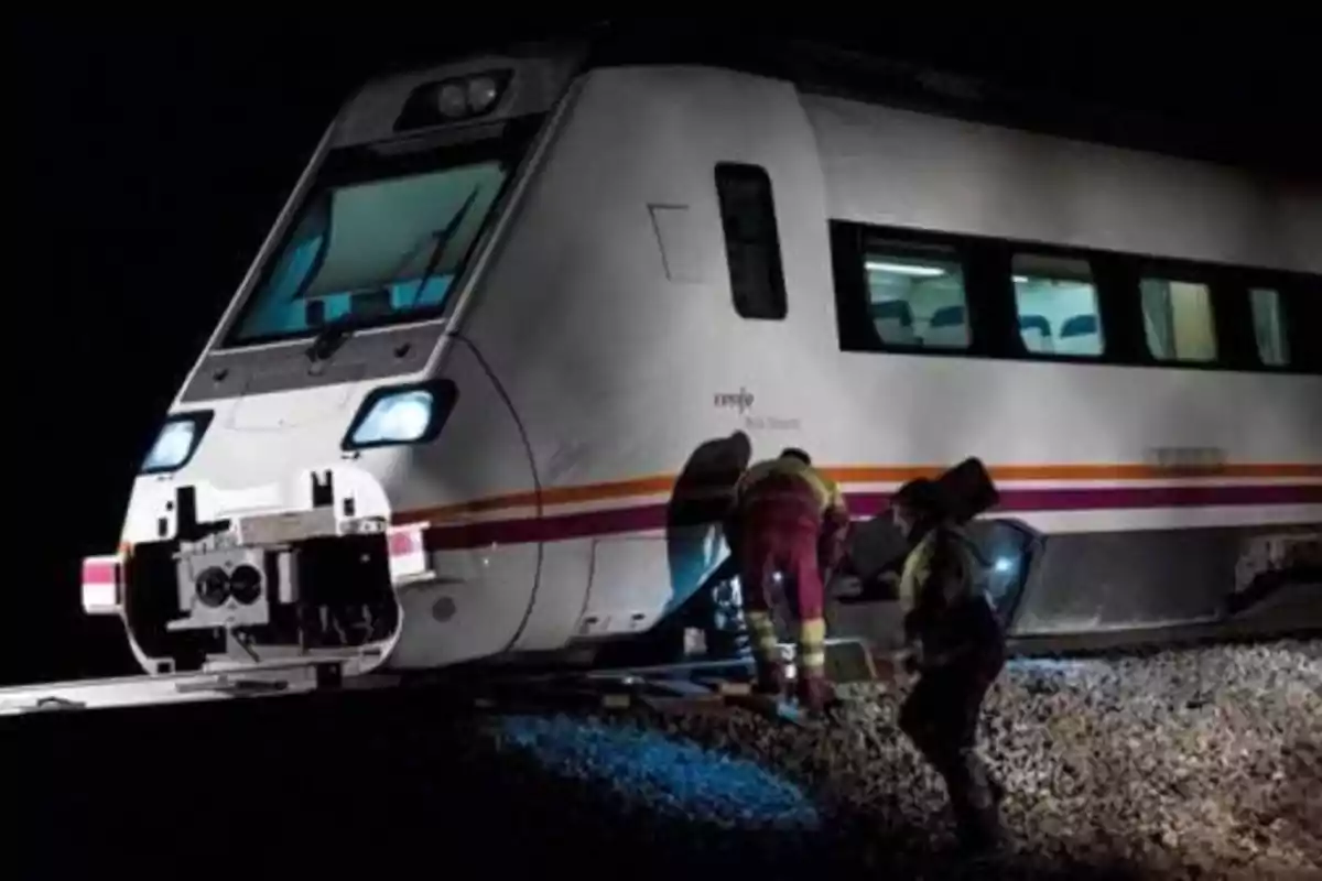 Un tren detenido en las vías durante la noche con dos personas trabajando cerca de él.