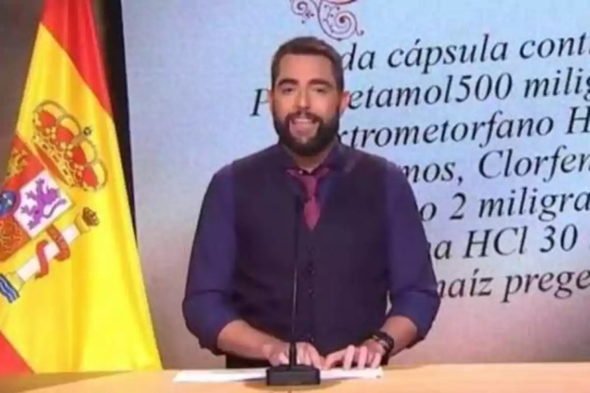 Un hombre de pie frente a un micrófono con una bandera de España a su izquierda y un texto en la pantalla detrás de él.