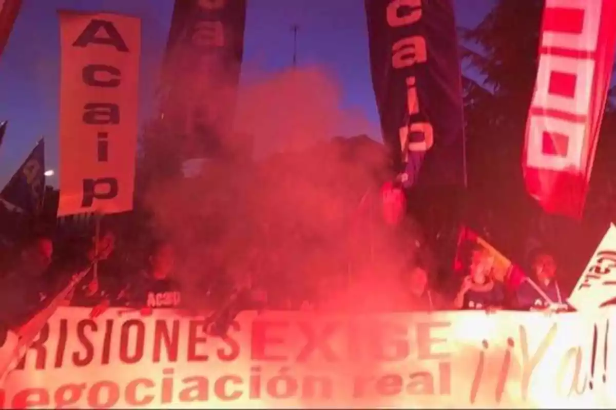 Manifestación nocturna con pancartas y humo rojo, donde se leen las palabras "Acaip" y "PRISIONES EXIGE negociación real ¡¡Ya!!".