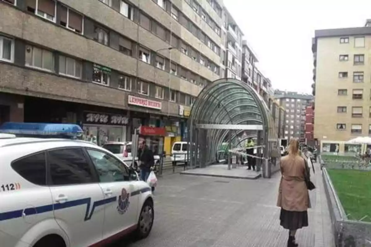 Una calle con edificios altos, una entrada de metro con estructura de vidrio y un coche de policía estacionado.