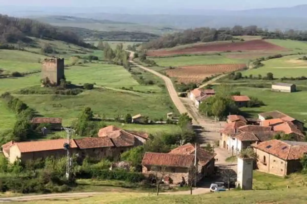 Vista panorámica de un pequeño pueblo rural con casas de tejados rojos, una torre antigua y campos verdes y marrones en el fondo.