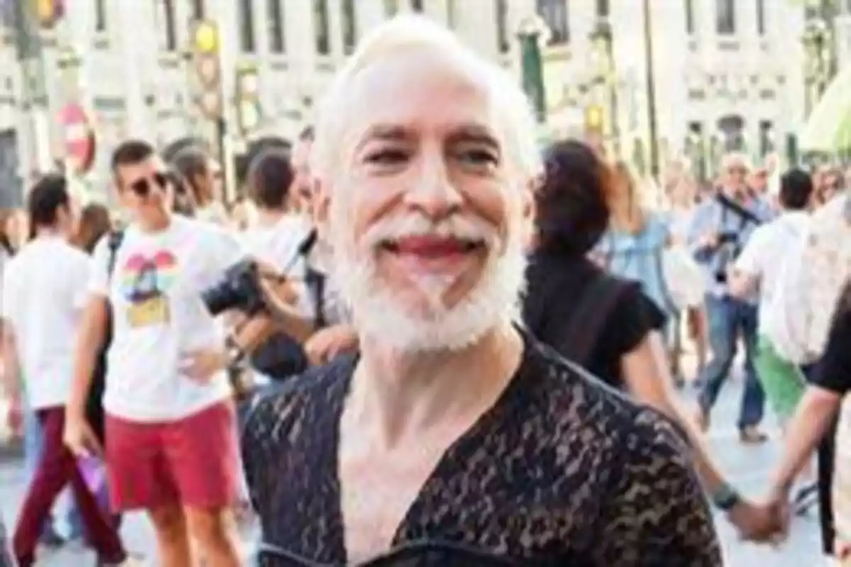 Una persona con barba blanca y cabello blanco sonríe mientras está rodeada de una multitud en una calle concurrida.