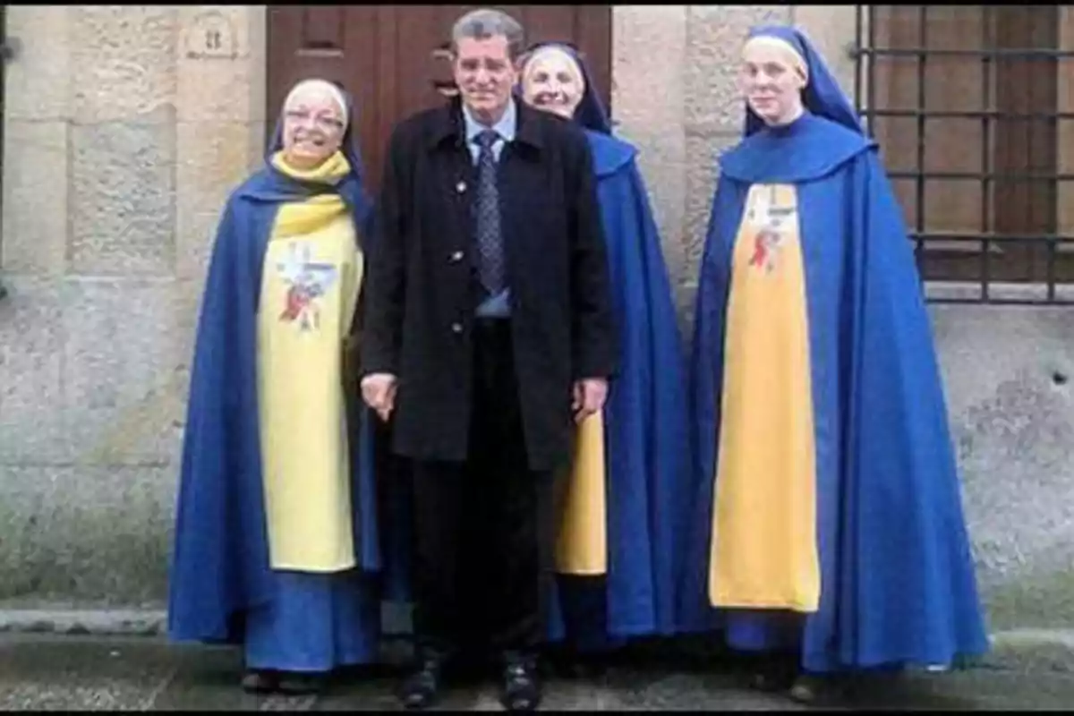 Un hombre de traje oscuro posa junto a tres monjas con hábitos azules y amarillos frente a una puerta de madera.