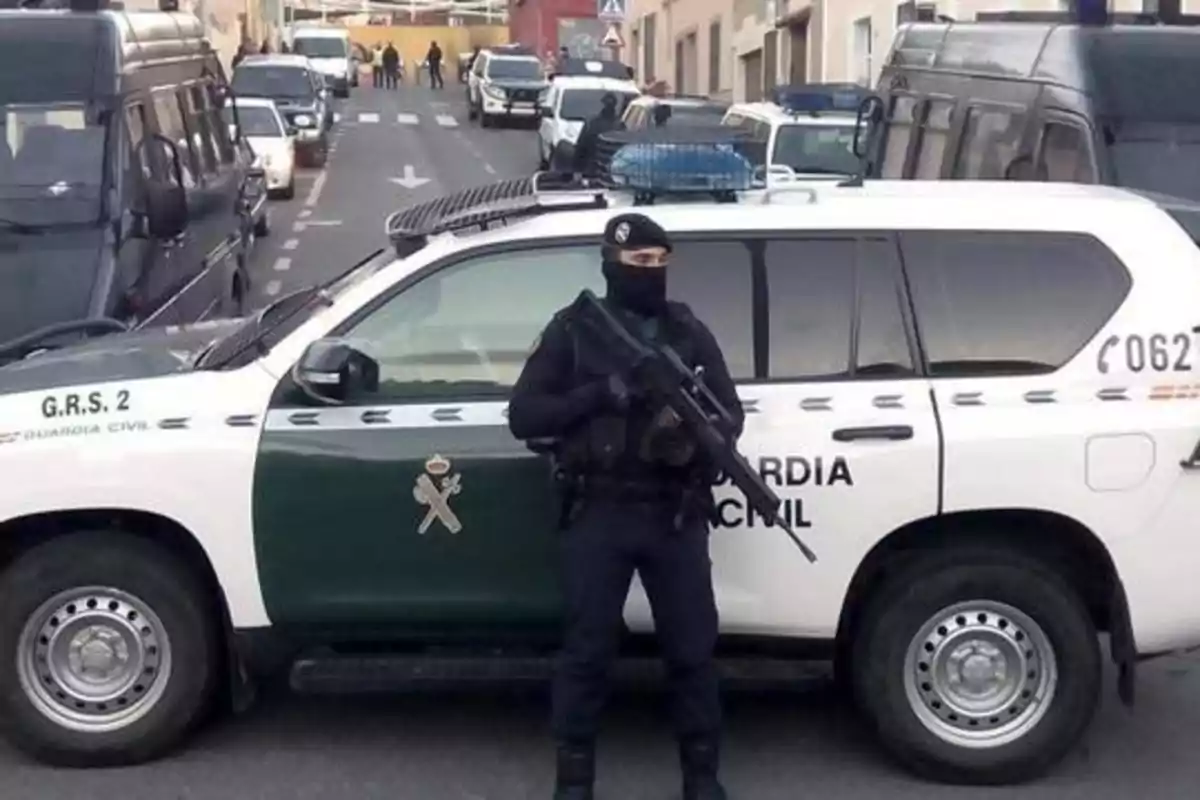 Un agente de la Guardia Civil armado y enmascarado se encuentra de pie frente a un vehículo oficial en una calle con varios vehículos estacionados y personas al fondo.