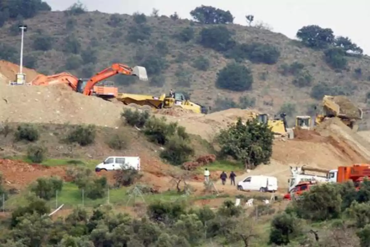 Excavadoras y maquinaria pesada trabajando en una obra de construcción en una zona montañosa con vegetación y varios vehículos estacionados.