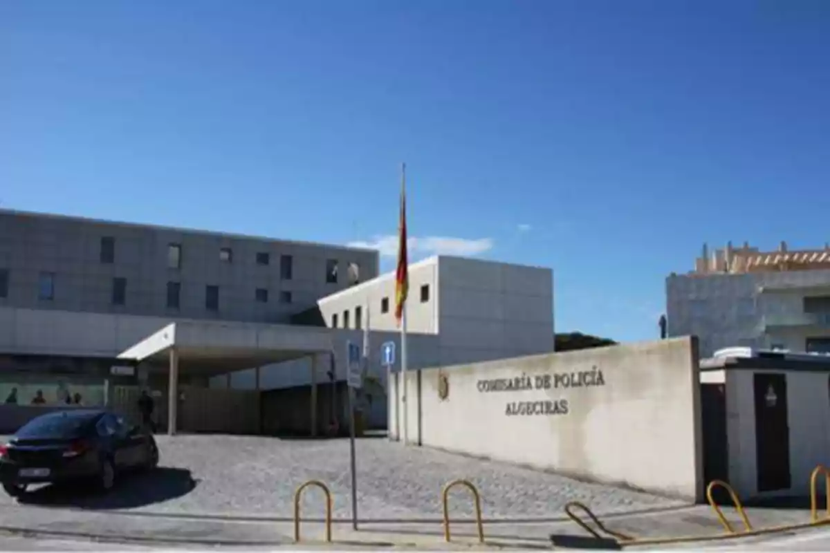 Edificio de la Comisaría de Policía de Algeciras con una bandera ondeando en el centro y un coche estacionado en la entrada.