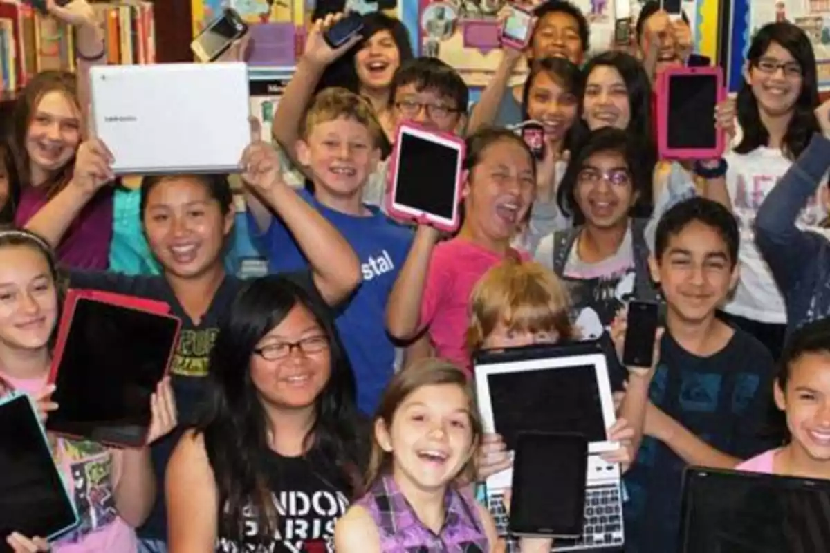 Un grupo de niños sonrientes sosteniendo dispositivos electrónicos como tabletas y teléfonos móviles en un entorno escolar.