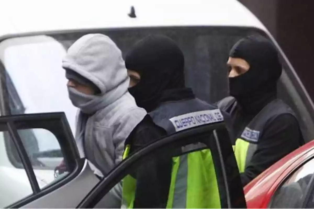 Agentes de policía con pasamontañas escoltan a una persona con capucha y bufanda hacia un vehículo.