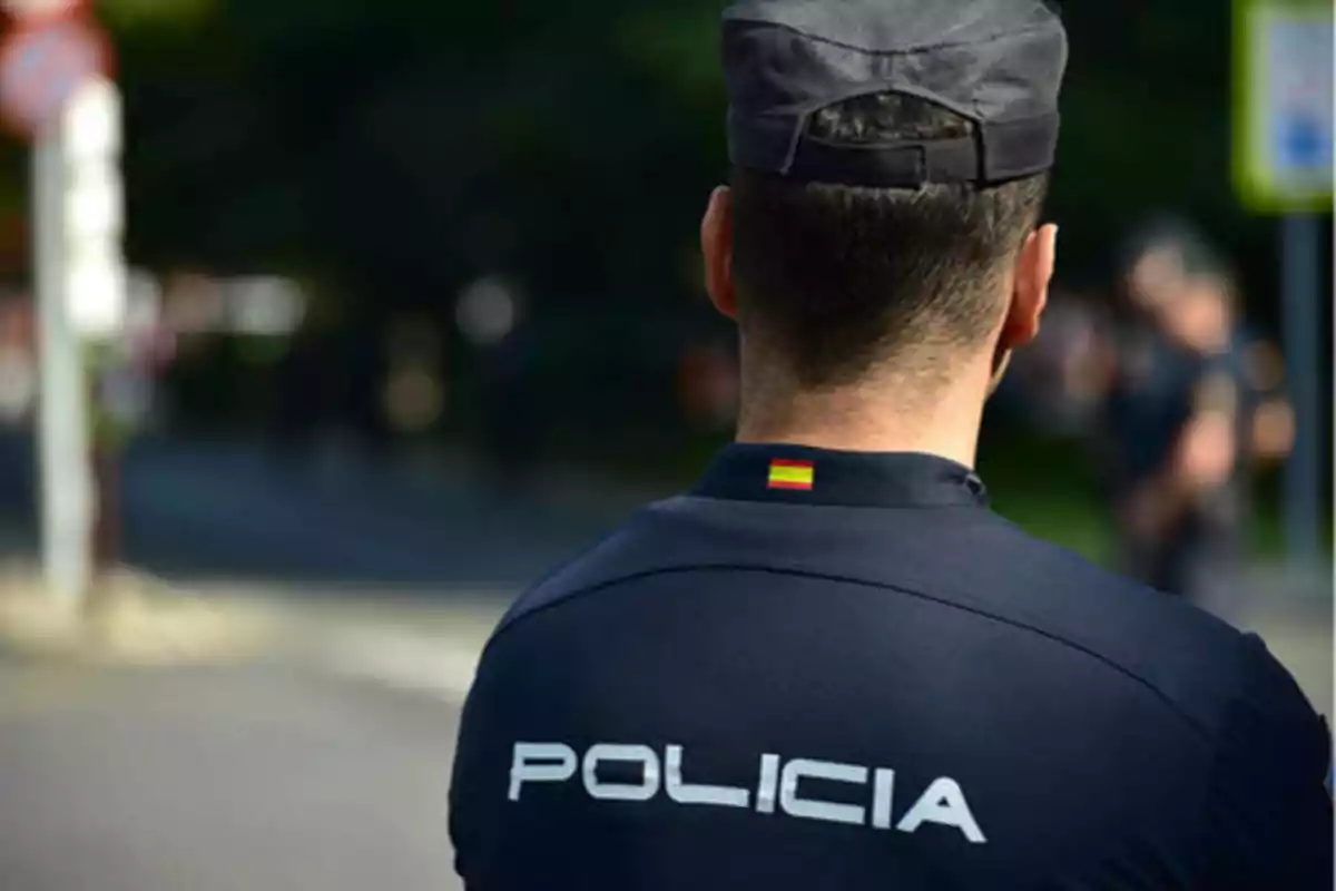 Un policía de espaldas con uniforme azul oscuro y una gorra, con la palabra "POLICÍA" escrita en la parte posterior de su chaqueta.