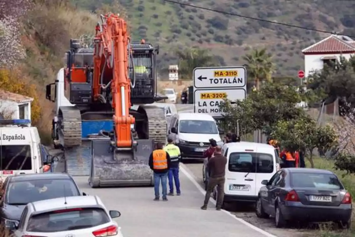 Una excavadora grande bloquea una carretera rural mientras varias personas y vehículos están detenidos alrededor, con señales que indican direcciones hacia Totalán y La Cala del Moral.