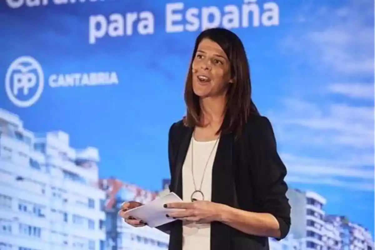 Una mujer hablando en un evento del Partido Popular en Cantabria con un fondo de edificios y un cielo azul.
