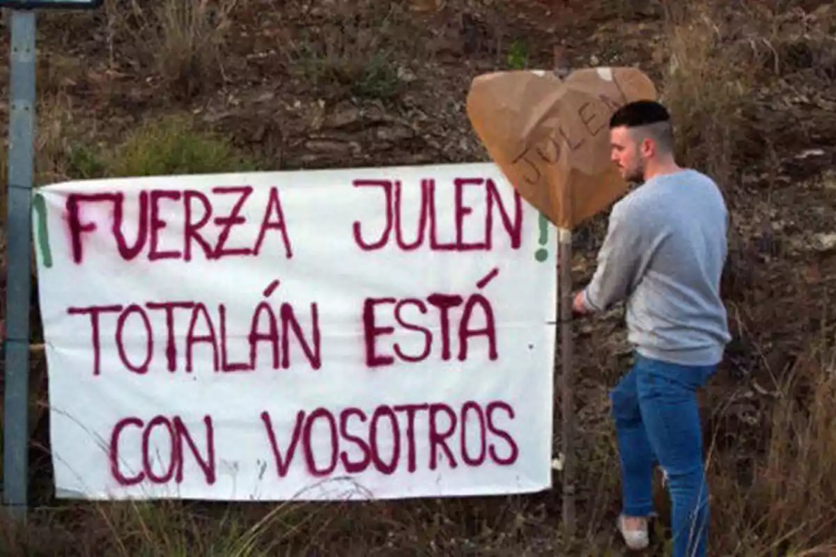 Un hombre coloca un cartel que dice "Fuerza Julen, Totalán está con vosotros" junto a un corazón de papel marrón con el nombre "Julen" escrito en él.