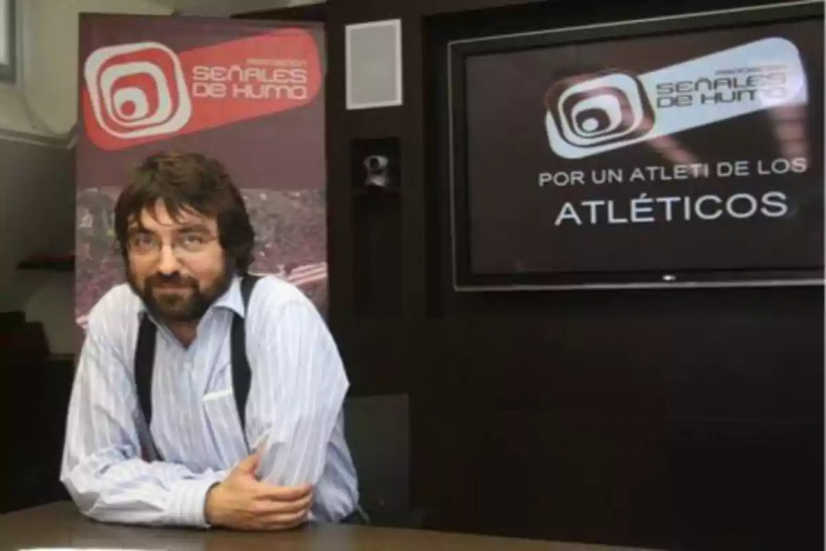 Un hombre con barba y gafas está sentado frente a una mesa, con un cartel de "Señales de Humo" y una pantalla que dice "Por un Atleti de los Atléticos" detrás de él.