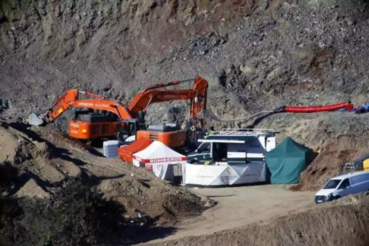 Excavadoras naranjas trabajando en una zona montañosa con tiendas de campaña y vehículos de apoyo.