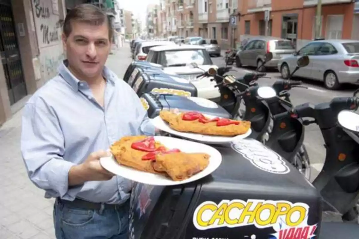 Un hombre sostiene dos platos con cachopos en una calle con varias motocicletas estacionadas.