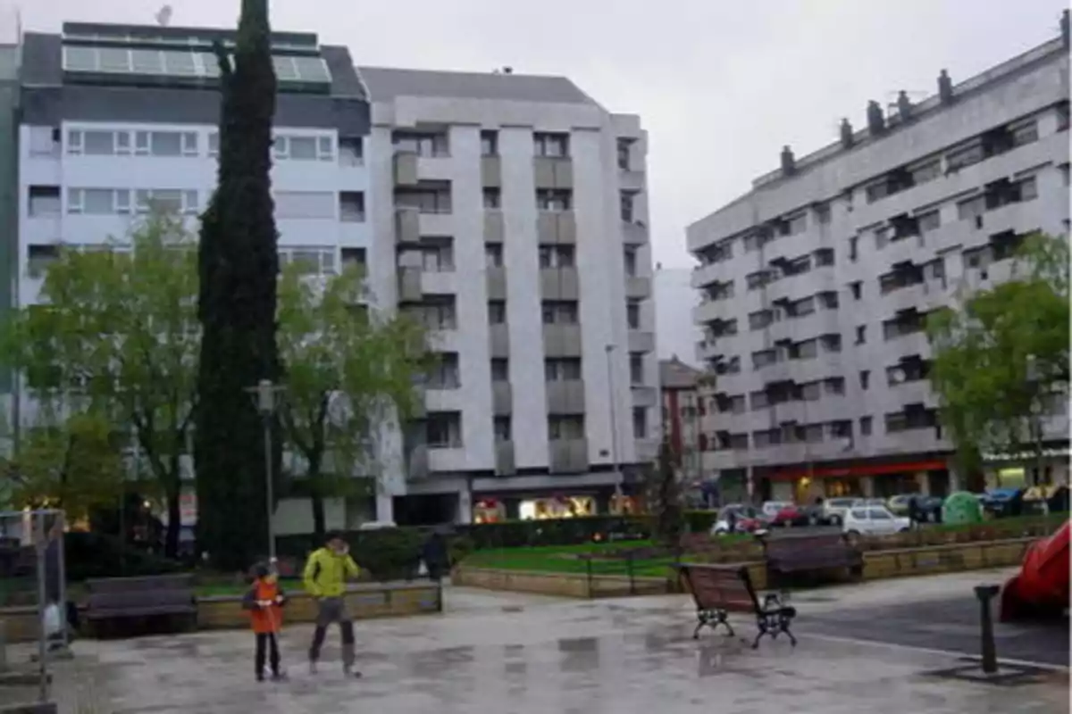 Una plaza con bancos y personas caminando, rodeada de edificios altos y árboles.