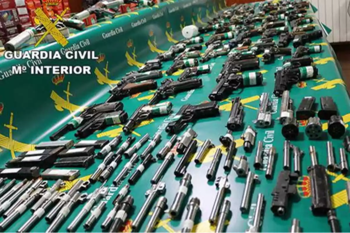 Una mesa verde con el logotipo de la Guardia Civil está cubierta de varias armas de fuego y piezas de armas desmanteladas.
