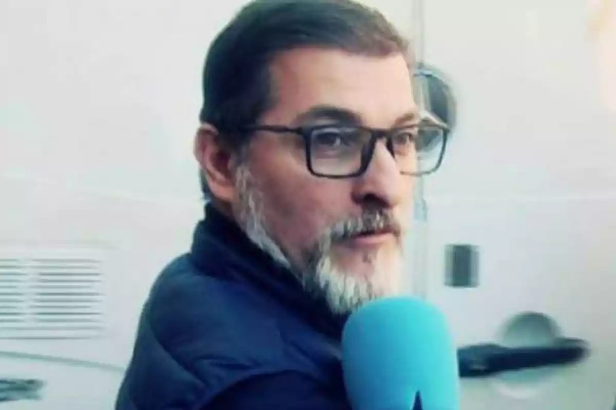Hombre con gafas y barba sosteniendo un micrófono azul.