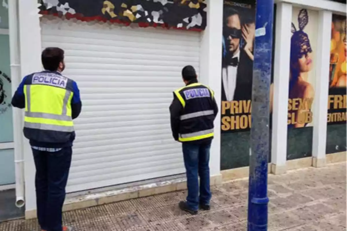 Dos policías de espaldas frente a un local con la persiana cerrada y carteles de un club privado en la fachada.