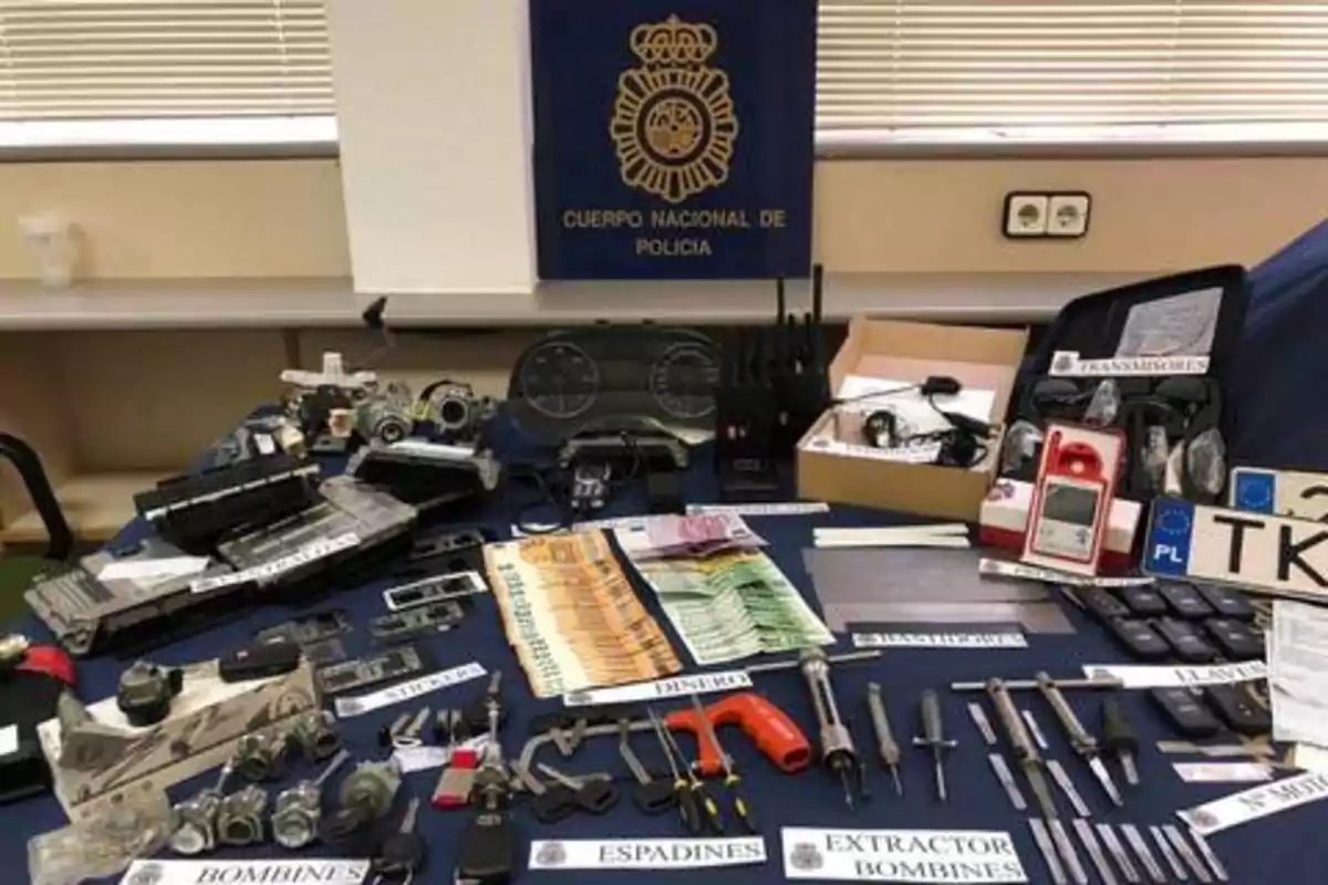 Mesa con diversos objetos incautados por la policía, incluyendo herramientas, dinero en efectivo, dispositivos electrónicos y matrículas de vehículos, con un cartel del Cuerpo Nacional de Policía en el fondo.