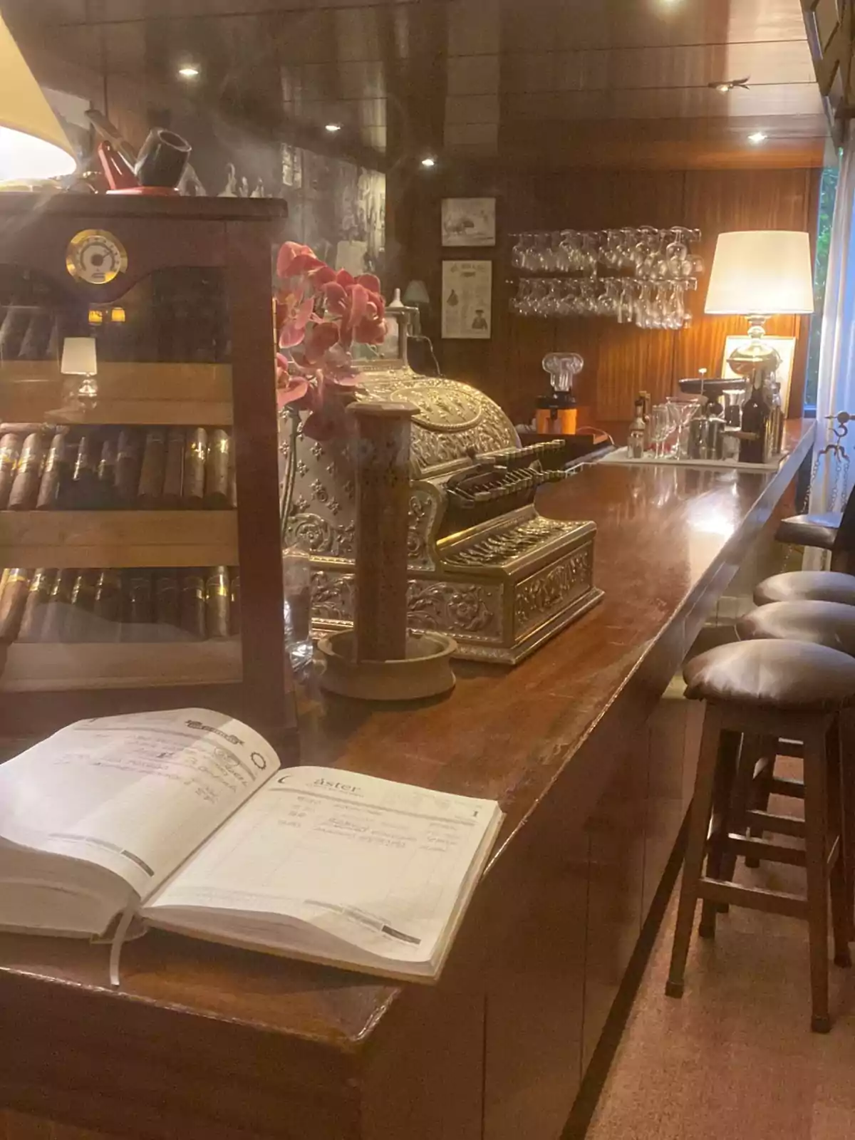 Una barra de bar con una caja registradora antigua, un libro abierto, una vitrina con puros y copas colgadas en la pared.