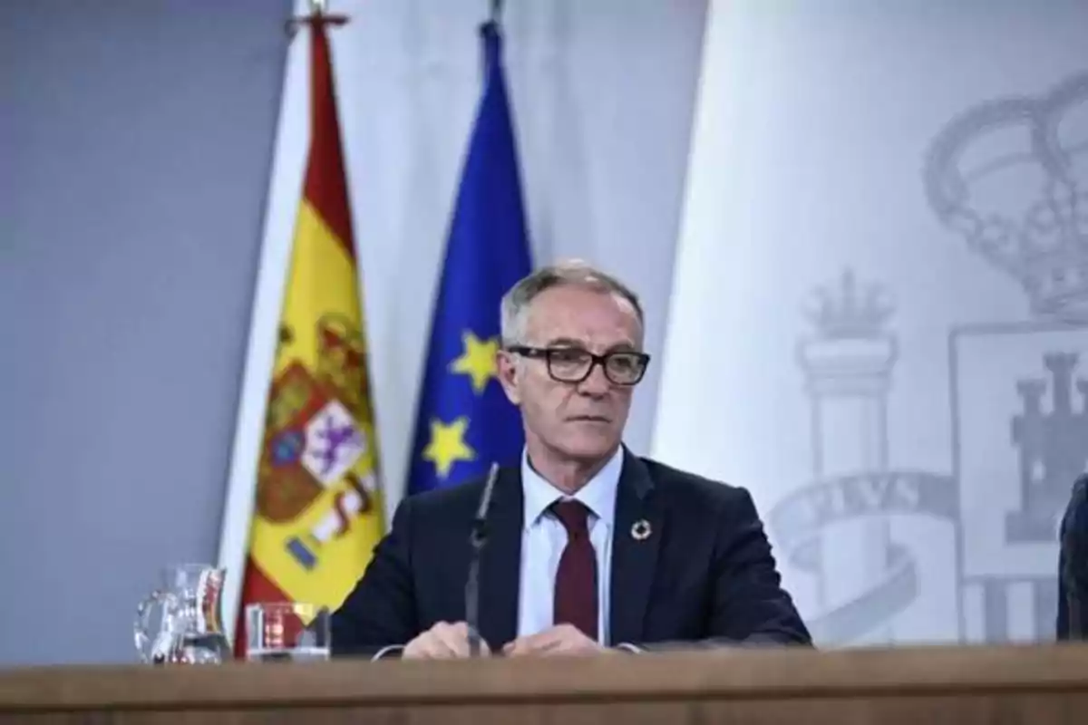 Hombre con gafas y traje oscuro sentado frente a micrófono con banderas de España y la Unión Europea de fondo.
