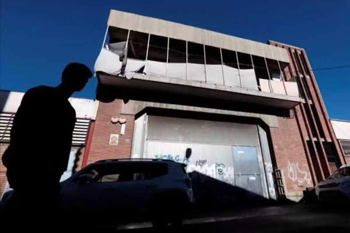 Un edificio de dos pisos con ventanas rotas y grafitis en la fachada, una persona en silueta y un coche estacionado en la calle.