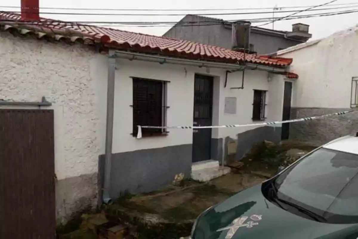 Una casa de una planta con techo de tejas rojas y paredes blancas, con ventanas enrejadas y una puerta de metal, está acordonada con una cinta de seguridad, y frente a ella hay un vehículo de la Guardia Civil.