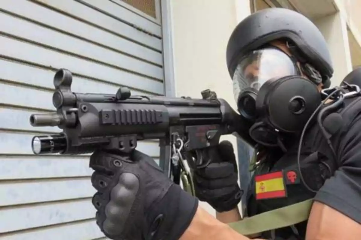 Un agente de fuerzas especiales con equipo táctico y máscara de gas apuntando con un arma frente a una pared metálica.