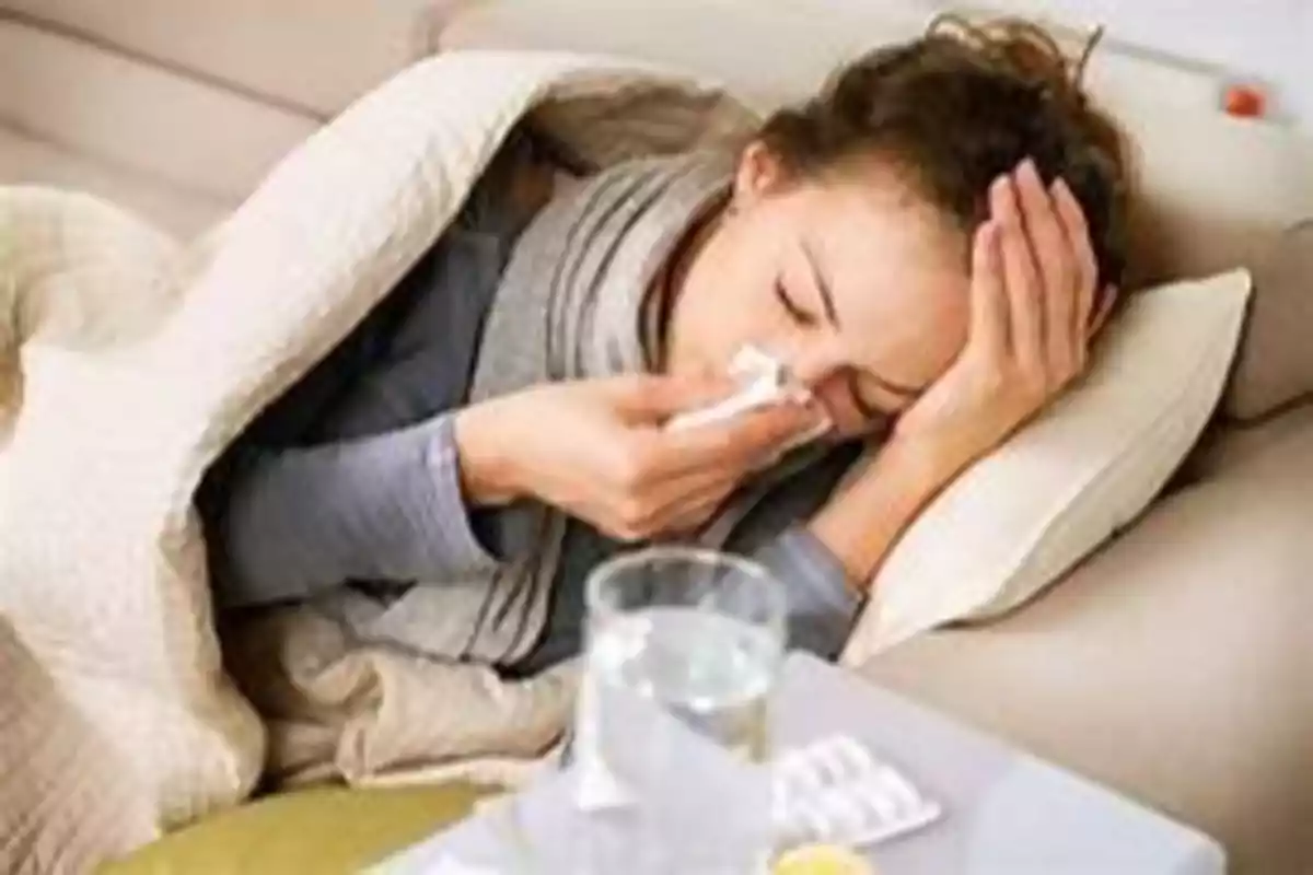 Persona enferma acostada en un sofá con una manta, usando un pañuelo para sonarse la nariz y con medicamentos y un vaso de agua en una mesa cercana.