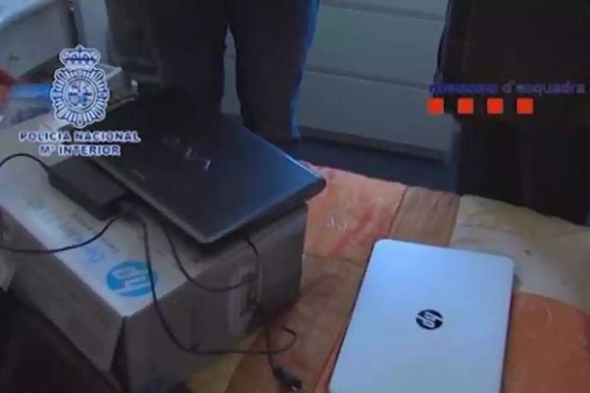 Dos computadoras portátiles sobre una cama, una de ellas conectada a un disco duro externo, con los logotipos de la Policía Nacional y los Mossos d'Esquadra en la imagen.