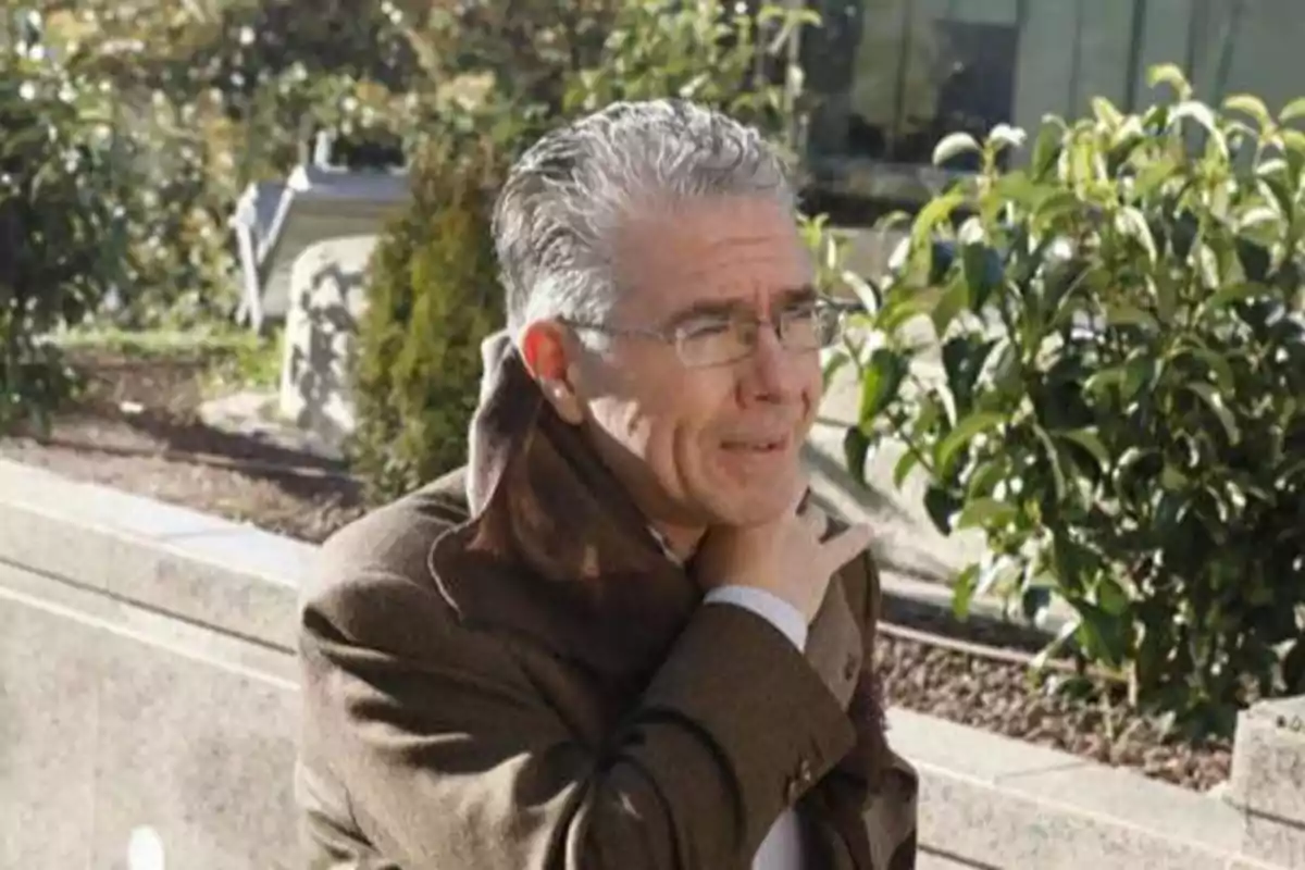 Un hombre con gafas y cabello canoso, vestido con un abrigo marrón, se encuentra al aire libre en un entorno urbano con vegetación al fondo.