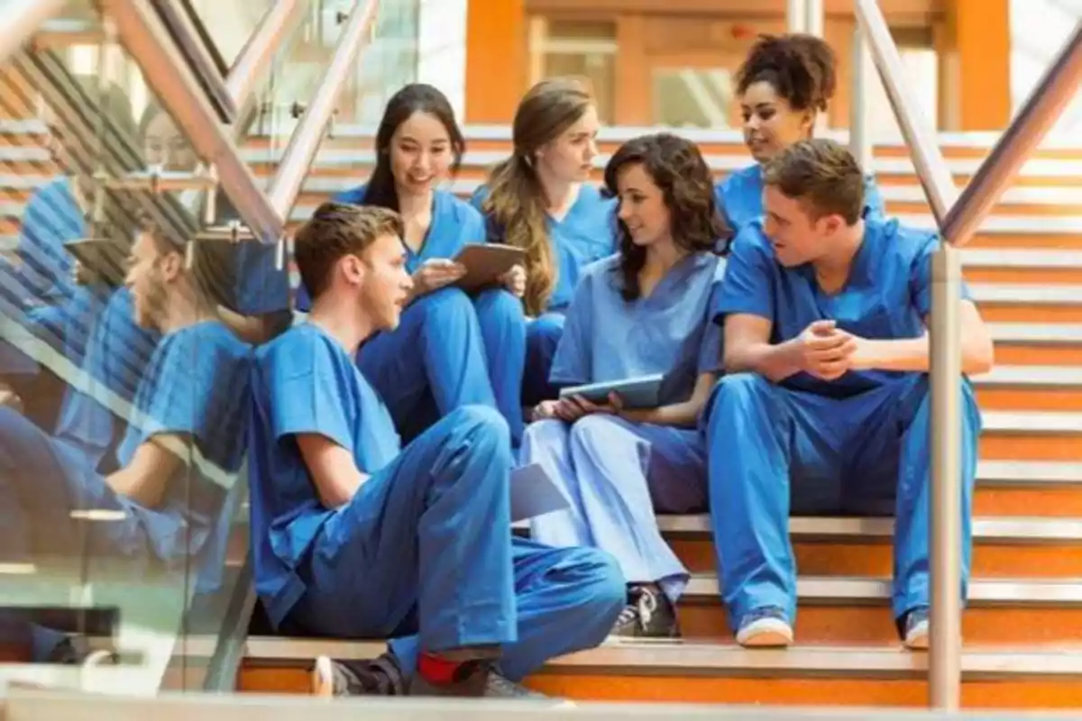 Un grupo de estudiantes de medicina con uniformes azules sentados en una escalera, conversando y usando tabletas.