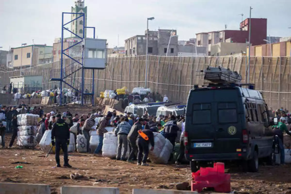 Personas trabajando y cargando bultos cerca de una valla fronteriza, con vehículos y edificios en el fondo.