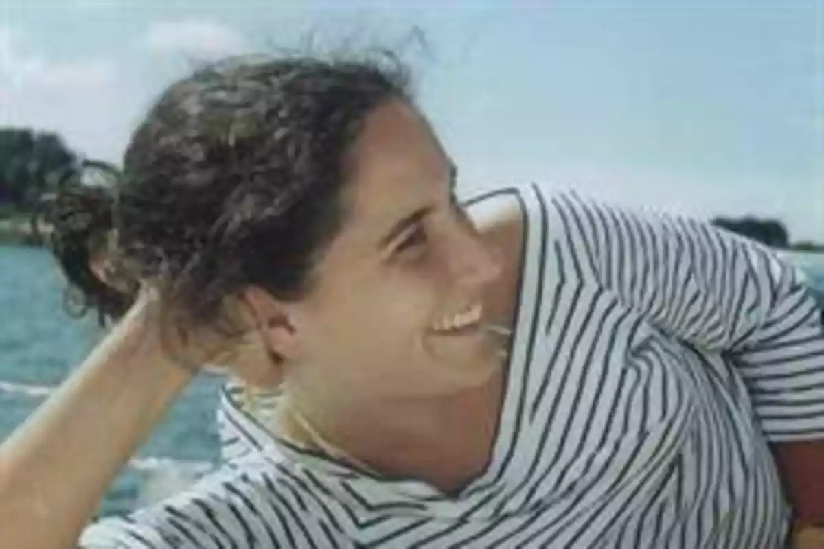 Persona sonriendo con una camiseta de rayas en un entorno al aire libre con agua de fondo.