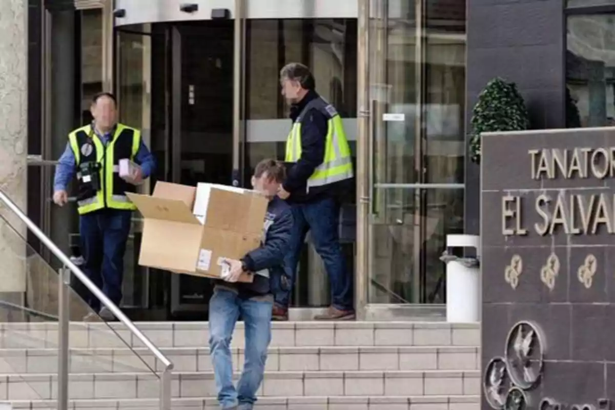 Tres personas con chalecos de seguridad llevan cajas y paquetes fuera de un edificio con un letrero que dice "Tanatorio El Salvador".