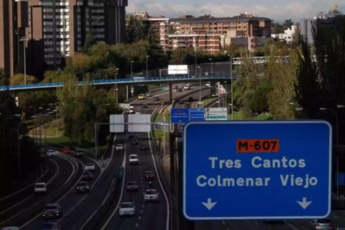 Una carretera con tráfico moderado, rodeada de edificios y vegetación, con un cartel que indica la dirección hacia Tres Cantos y Colmenar Viejo en la M-607.