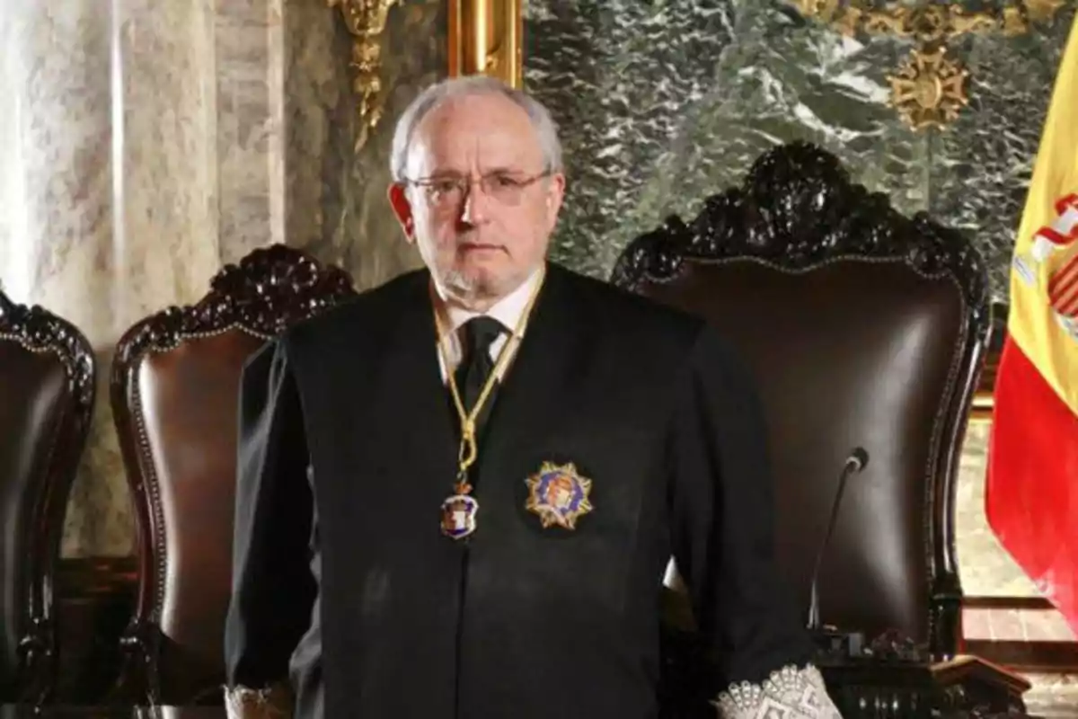 Un hombre mayor con gafas y cabello canoso, vestido con una toga negra y condecoraciones, de pie frente a un fondo de mármol y una bandera española.