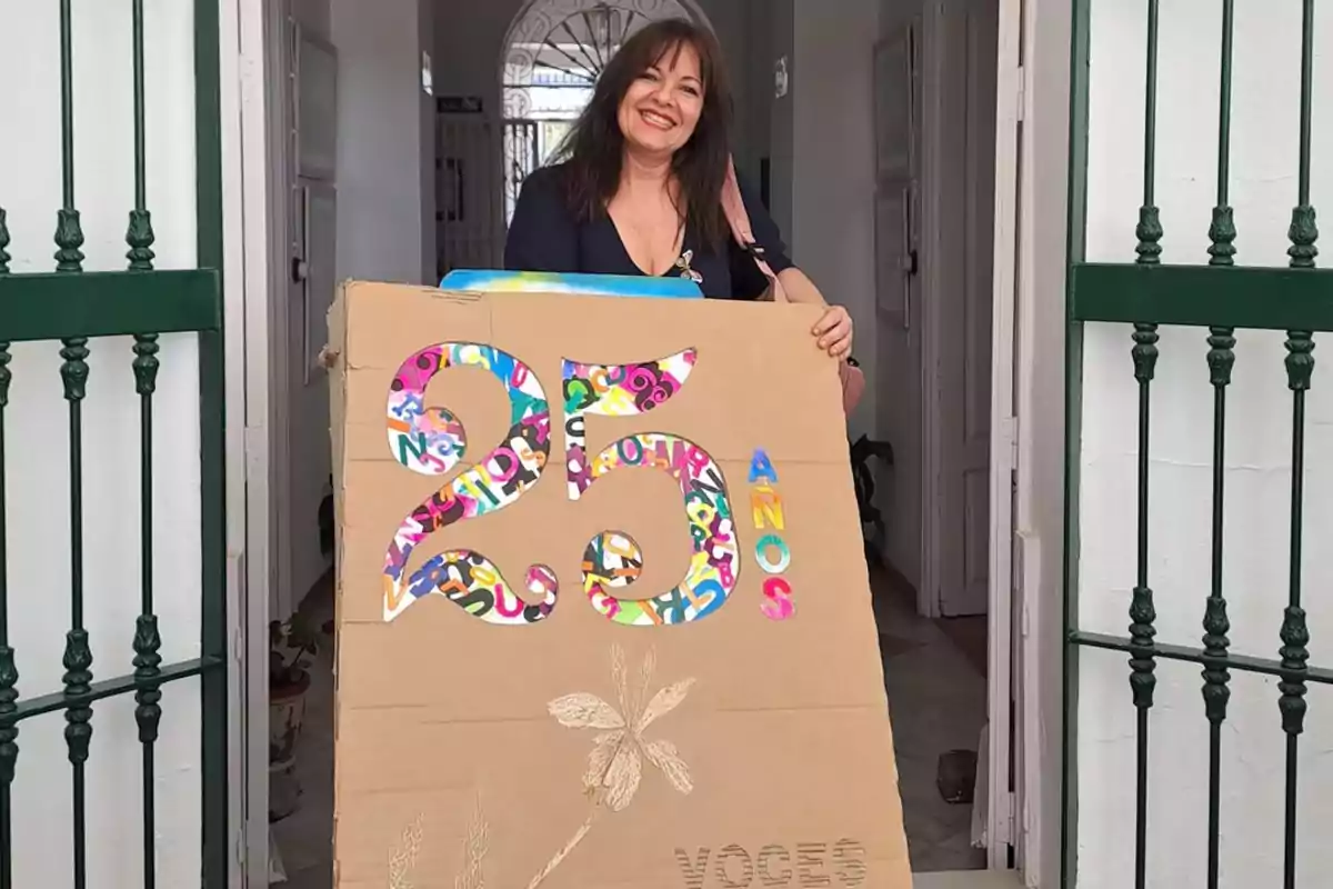 Una mujer sonriente sostiene un cartel de cartón que celebra 25 años con letras coloridas, de pie en la entrada de un edificio con puertas verdes.