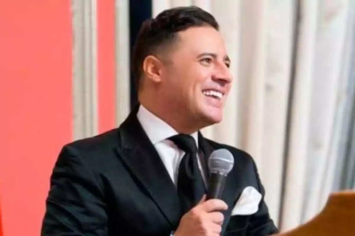 Hombre con traje negro y corbata sosteniendo un micrófono y sonriendo.