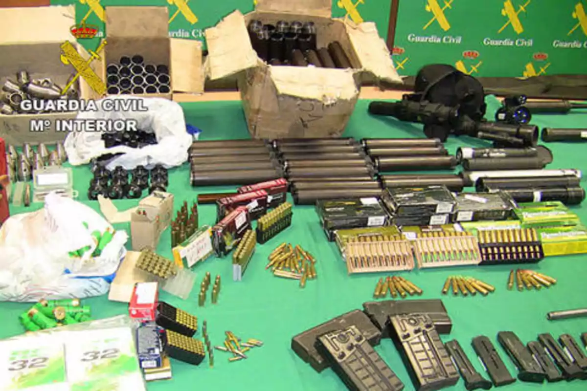 La imagen muestra una mesa con una gran cantidad de municiones, cargadores y otros componentes relacionados con armas, con un fondo que tiene el logotipo de la Guardia Civil y el Ministerio del Interior.