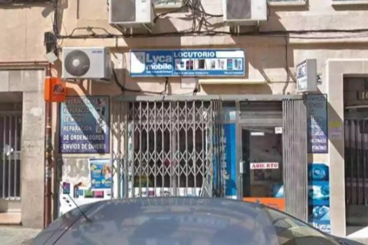La imagen muestra la fachada de un locutorio con varios carteles publicitarios, incluyendo uno de Lycamobile, y un coche estacionado frente a la entrada.