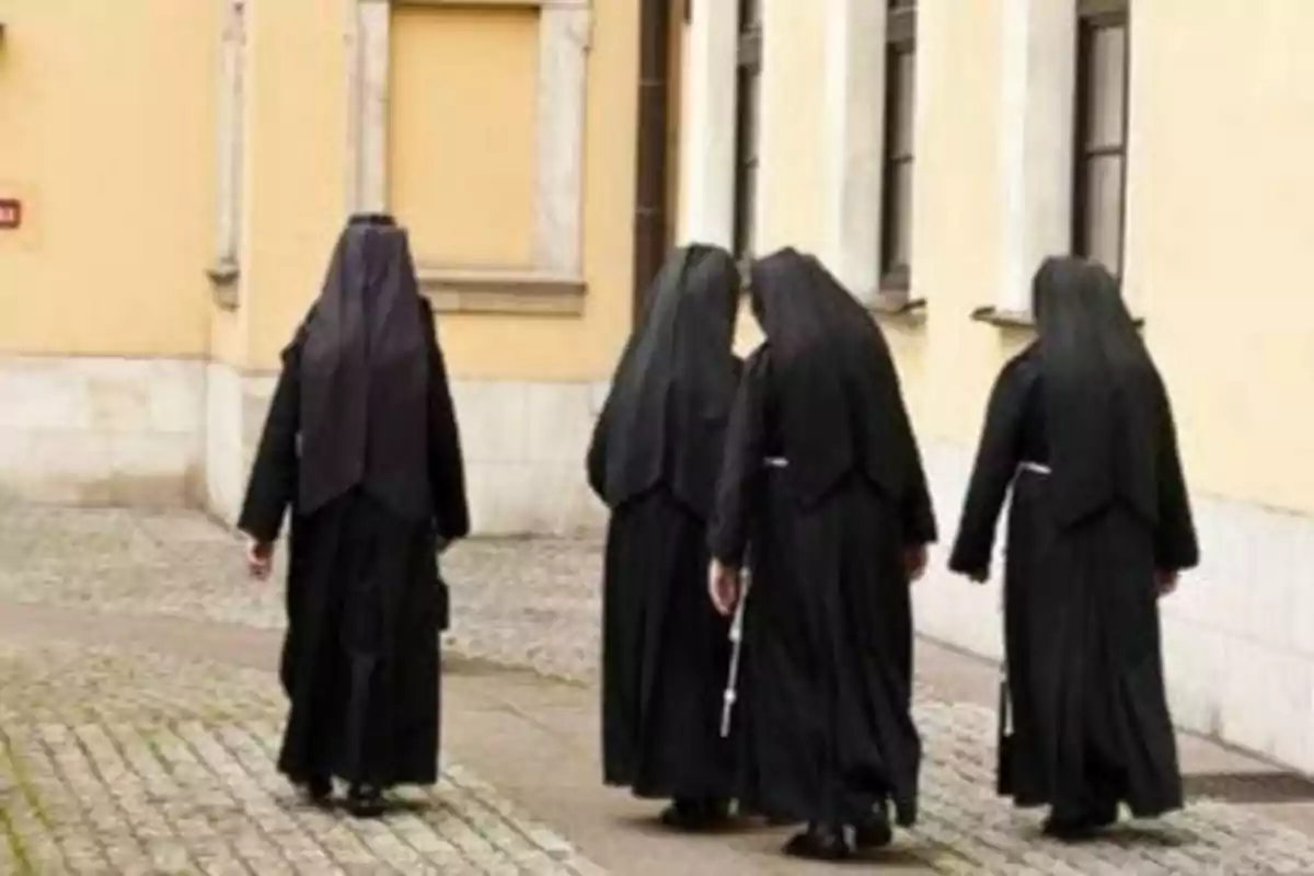 Cuatro monjas caminando por una calle empedrada junto a un edificio de paredes amarillas.