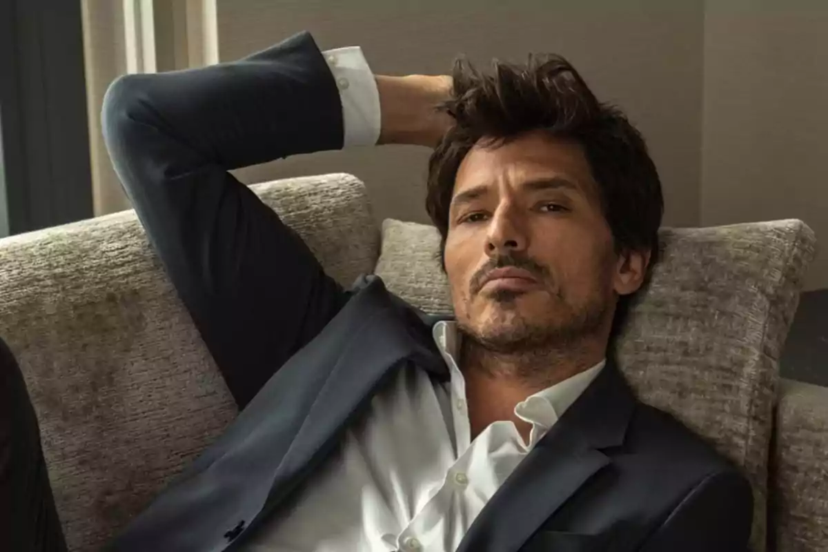 Un hombre con barba y cabello oscuro recostado en un sofá, vistiendo un traje oscuro y camisa blanca.