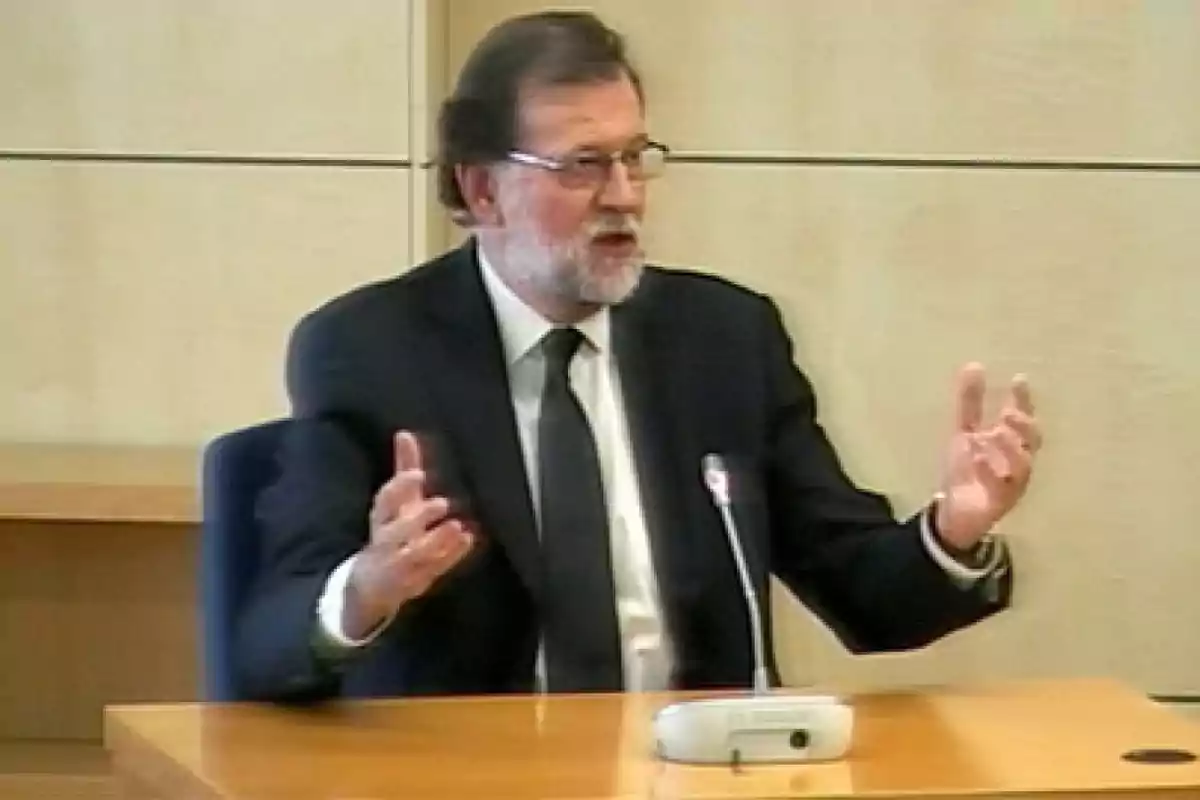 Hombre con barba y gafas, vestido con traje oscuro y corbata, hablando y gesticulando en una sala con un micrófono frente a él.
